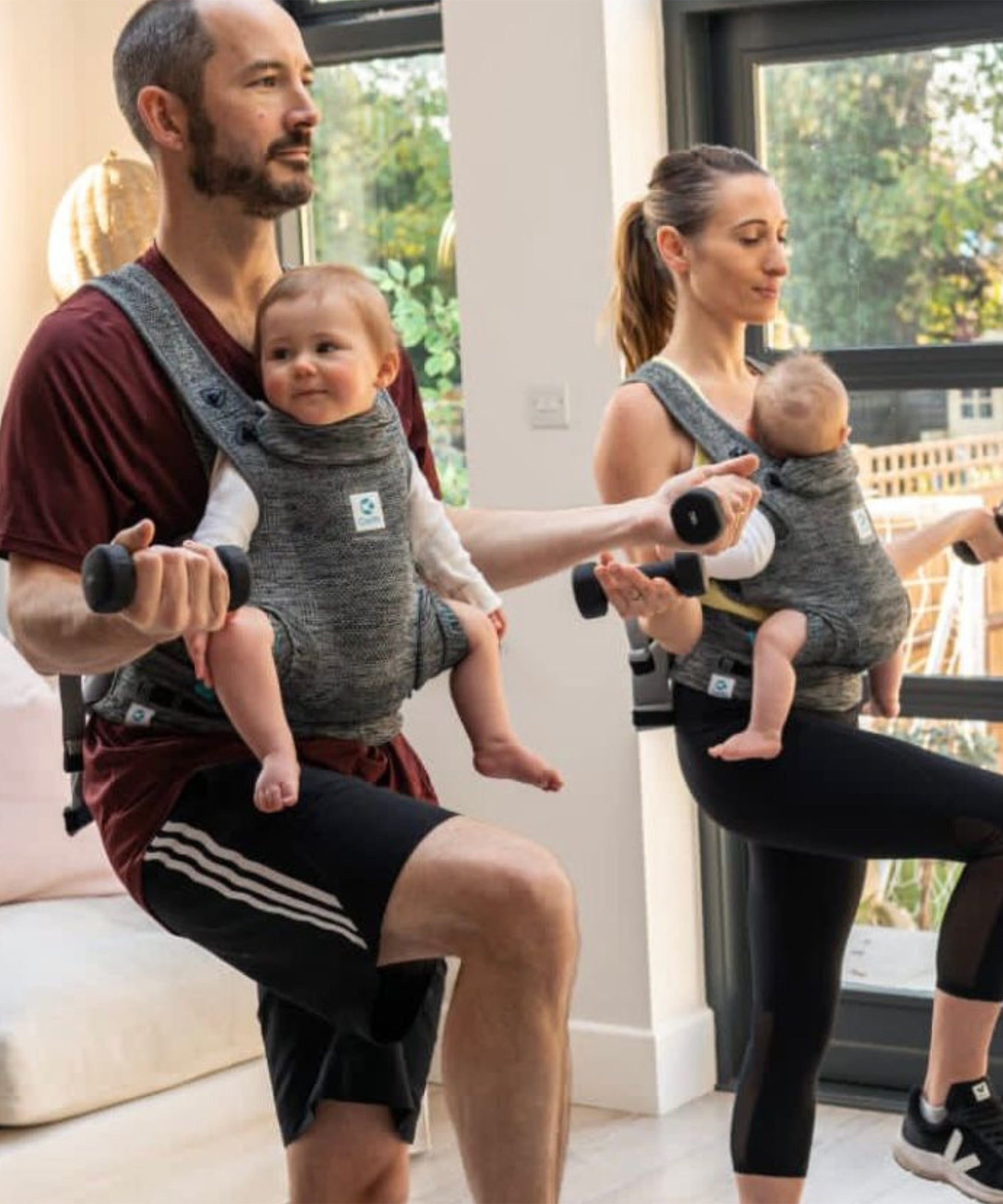 Two people working out and exercising at home, both carrying a child in a Carifit+ Baby Carrier in Cool Grey. The child on the left is outwards facing in the carrier, and the child on the right is inwards facing. The image shows how supported the child is inside the carrier and that parents can still exercise whist carrying their children in the carrier