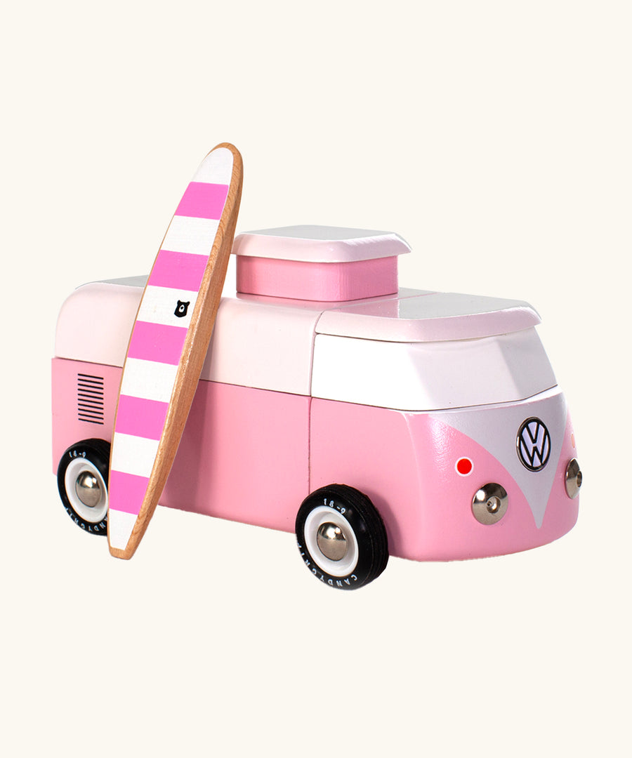 The Candylab VW Beach bus in pink, with a wooden pink and white stripe surf board propped up next to the van, The image is on a cream background