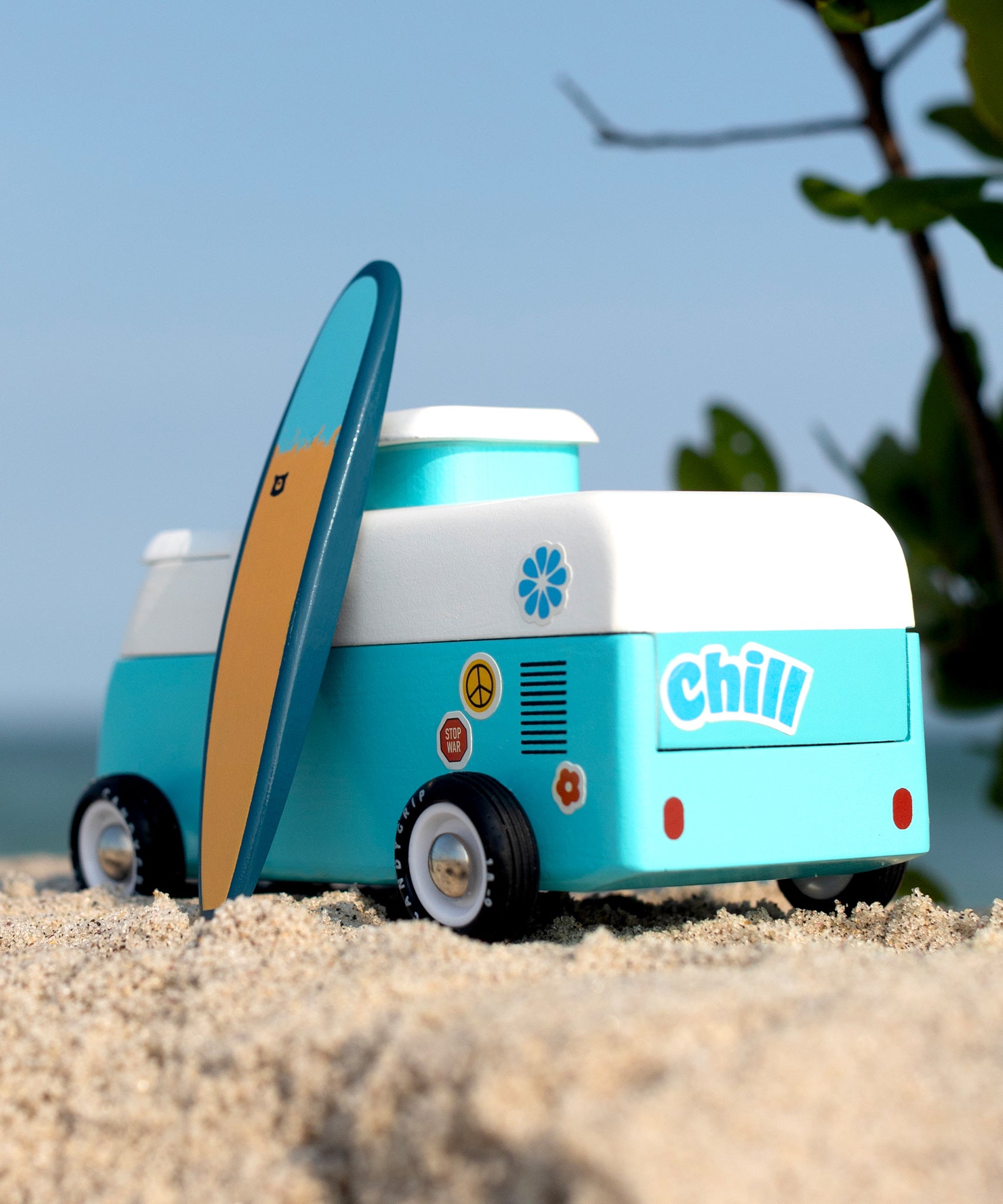 The back of the Candylab VW Beach bus in blue, with a blue and surf board propped up next to the van, stood on a sandy beach with a blue sky background. The image also shows colourful stickers on the back of the van