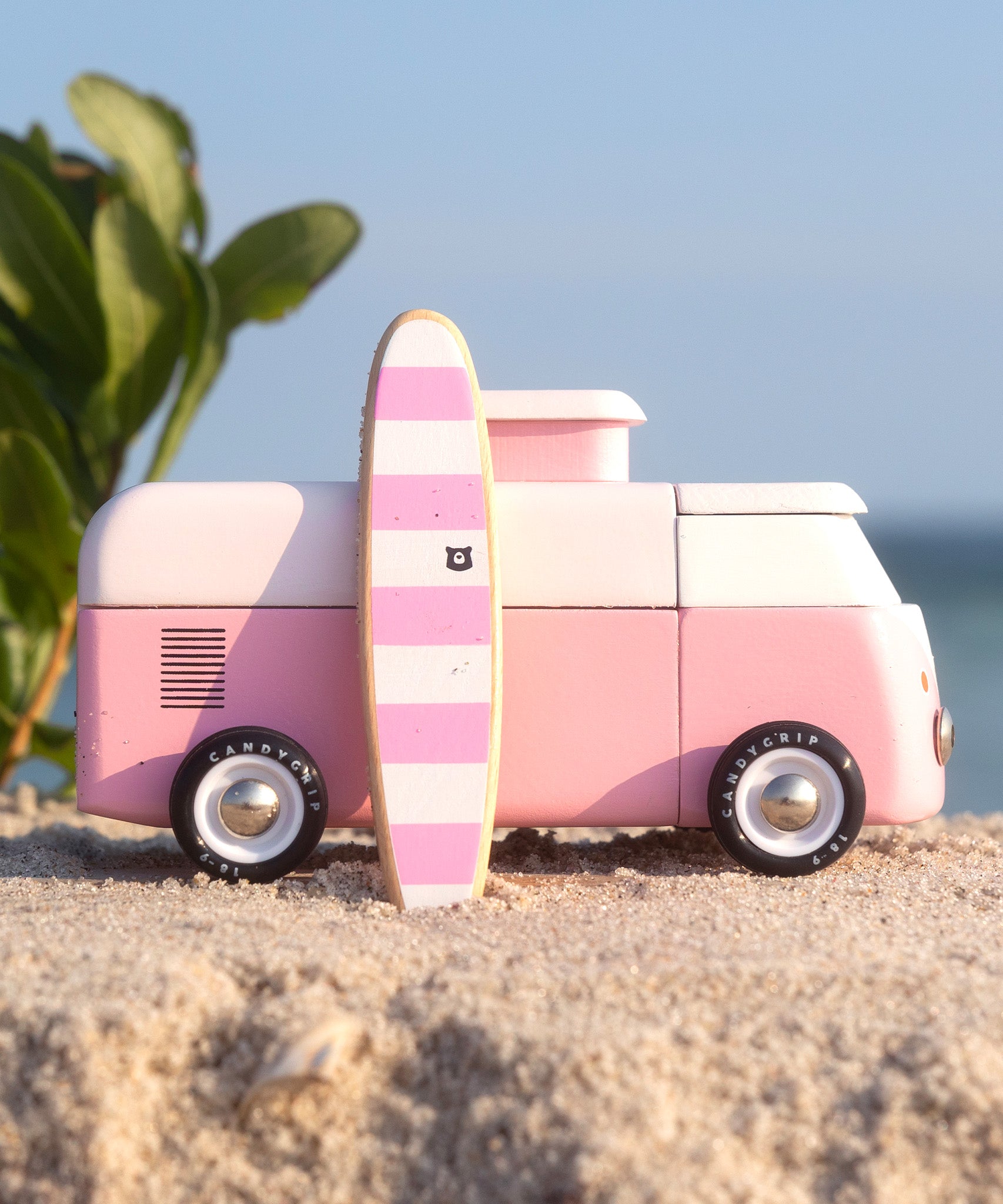 A side view of the Candylab VW Beach bus in pink, with a pink and white stripe surf board propped up next to the van, stood on a sandy beach with a blue sky background