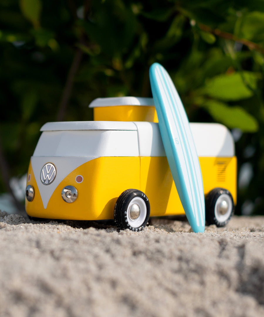 Candylab VW Beach bus in yellow, with a blue surf board propped up next to the van, stood on a sandy beach with green tree leaves and branches in the background