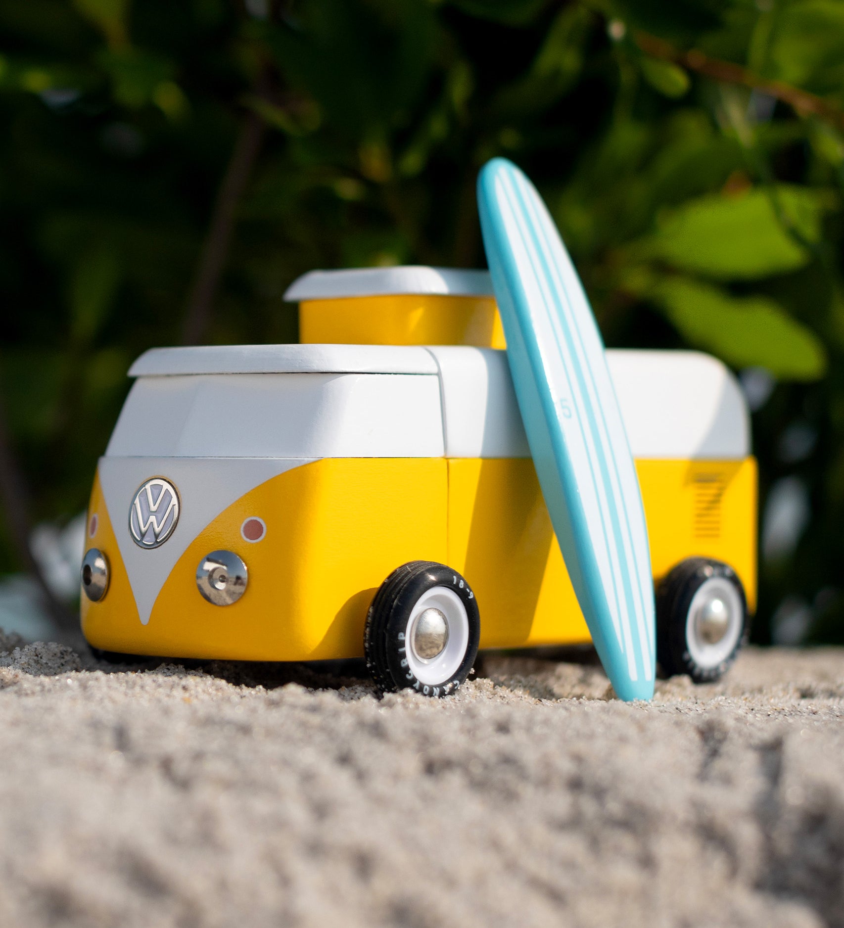 Candylab VW Beach bus in yellow, with a blue surf board propped up next to the van, stood on a sandy beach with green tree leaves and branches in the background