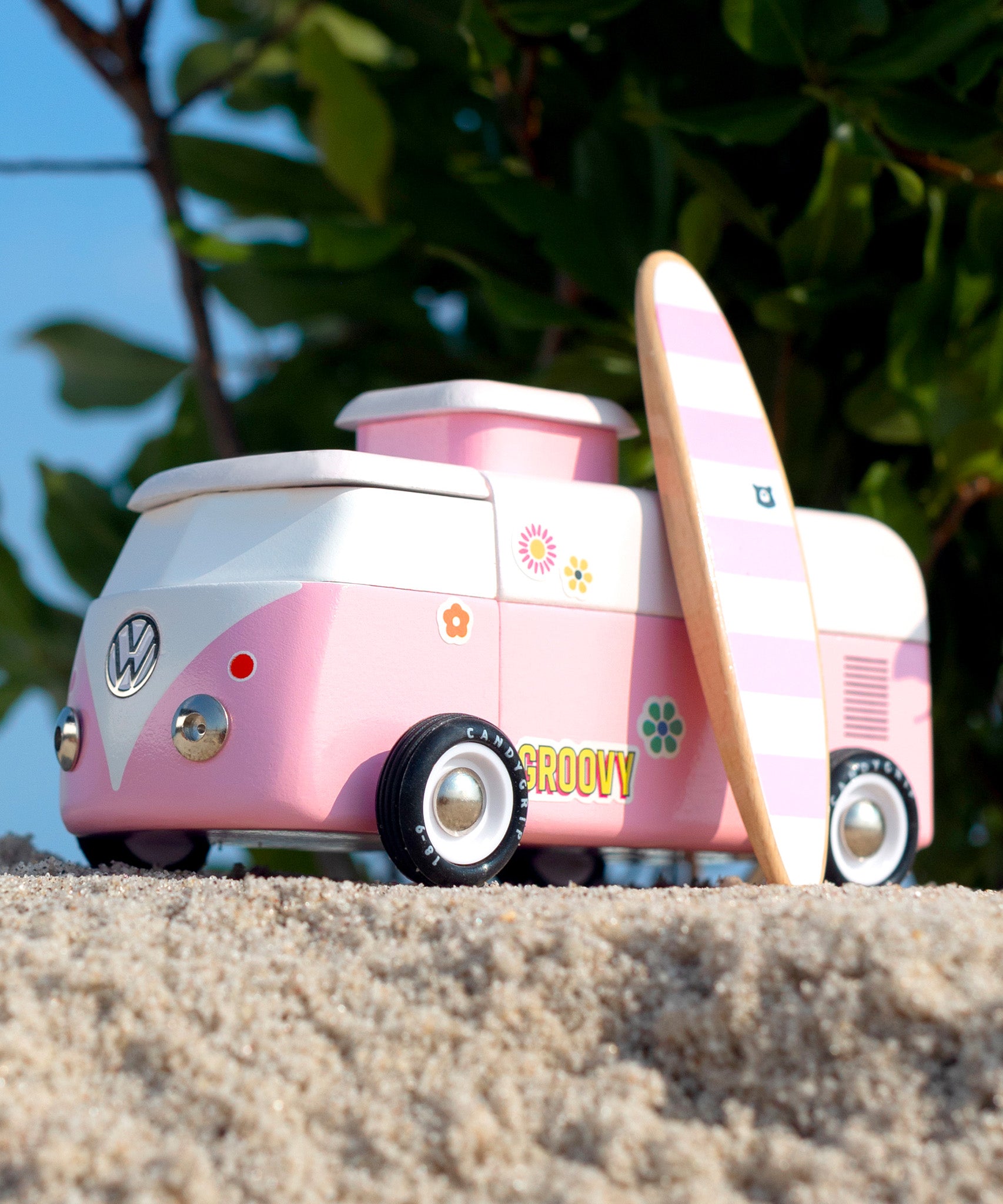 Candylab VW Beach bus in pink, with a pink and white stripe surf board propped up next to the van, stood on a sandy beach with green tree leaves and branches in the background