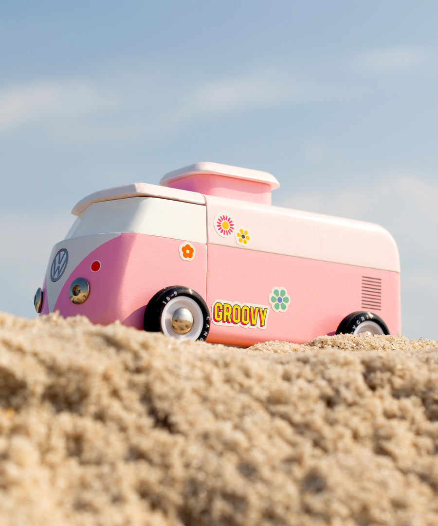 A side view of the Candylab VW Beach bus in pink with colourful stickers on the side of the van, stood on a sandy beach with a blue sky background