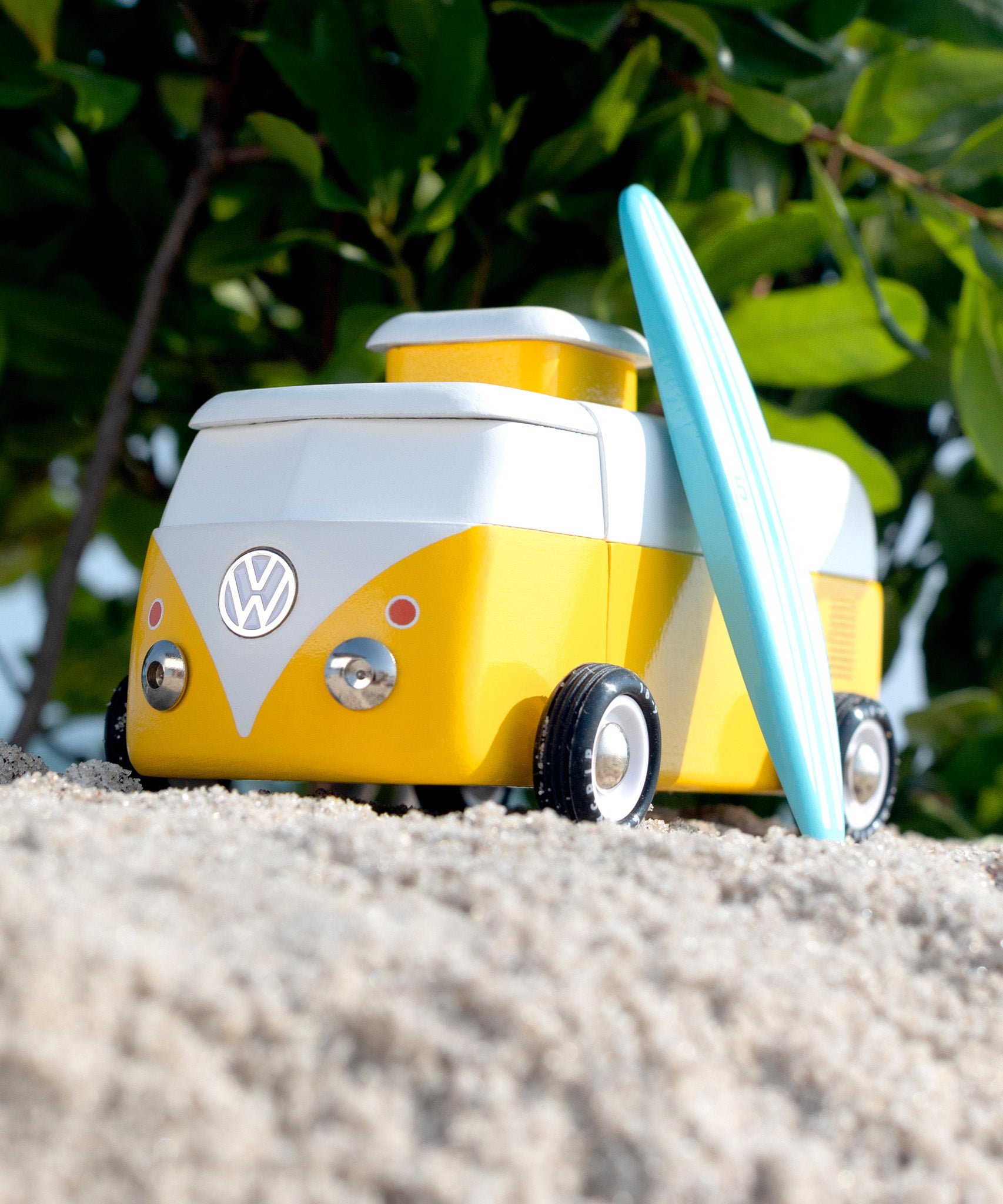 Candylab VW Beach bus in yellow, with a blue surf board propped up next to the van, stood on a sandy beach with green tree leaves and branches in the background. The image shows the front of the bus with a VW logo and silver studs to mimic head-lights