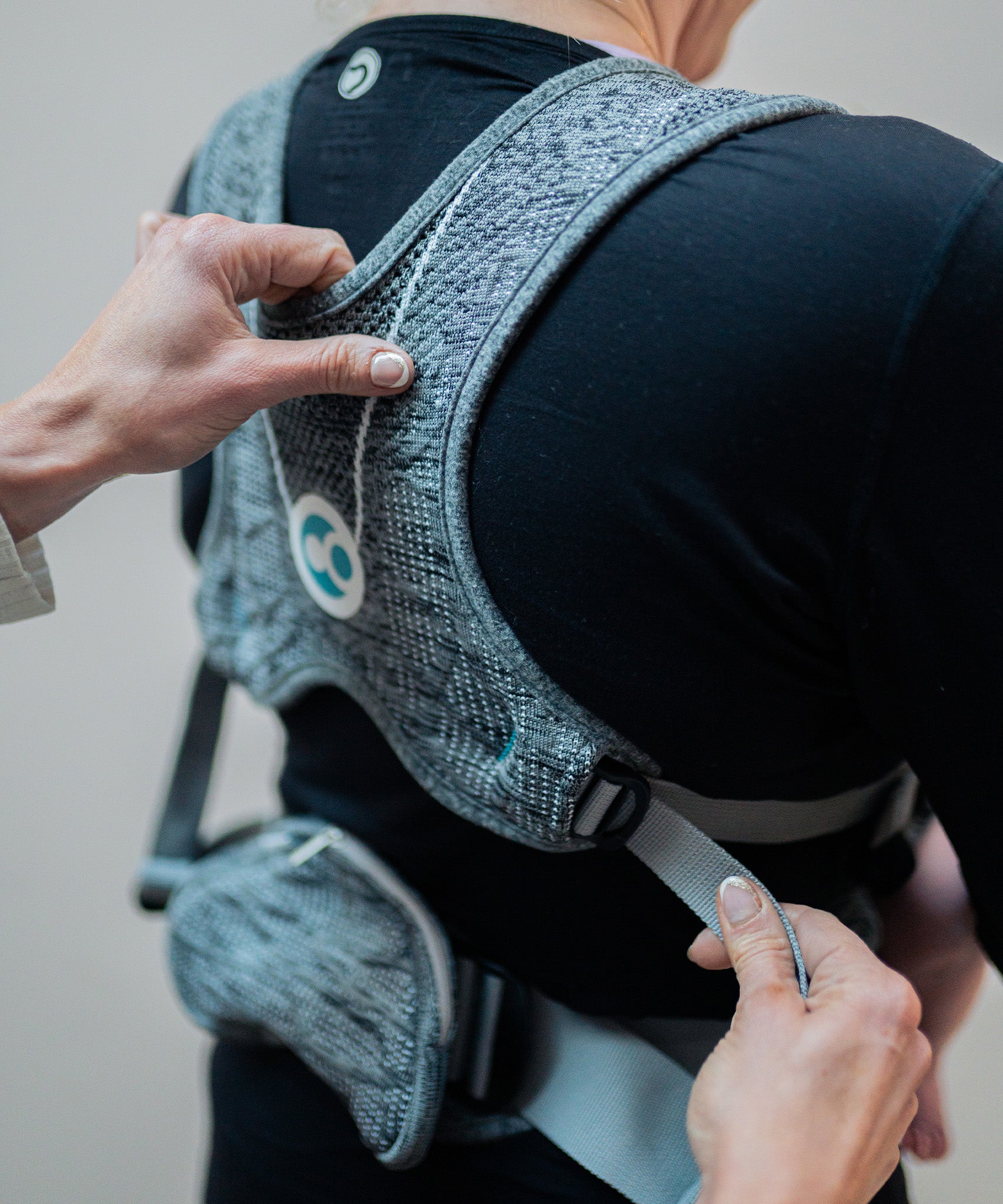 A person wearing the Carifit+ Baby Carrier in Cool Grey. The image shows the back on the carrier on the persons back, with another adult adjusting the adaptive sizing straps on the side