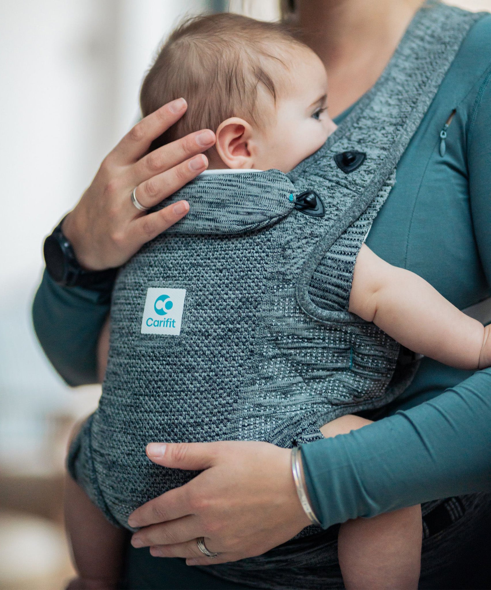 A child being carried in the Carifit+ Baby Carrier in Cool Grey. The child is facing inwards towards the adult