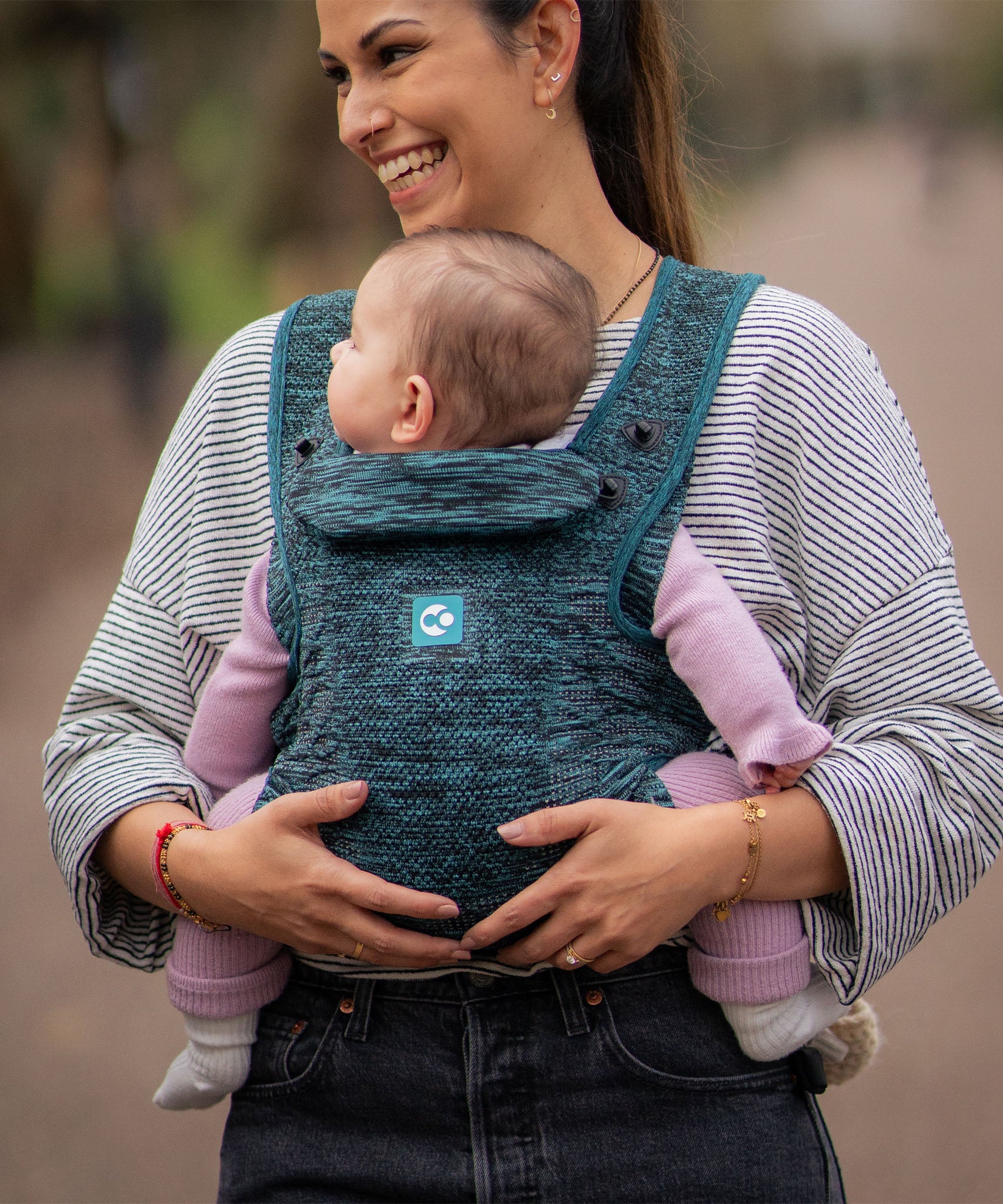 A person carrying a child in the Carifit+ Baby Carrier in Cool Green. The child is facing inwards towards the adult.