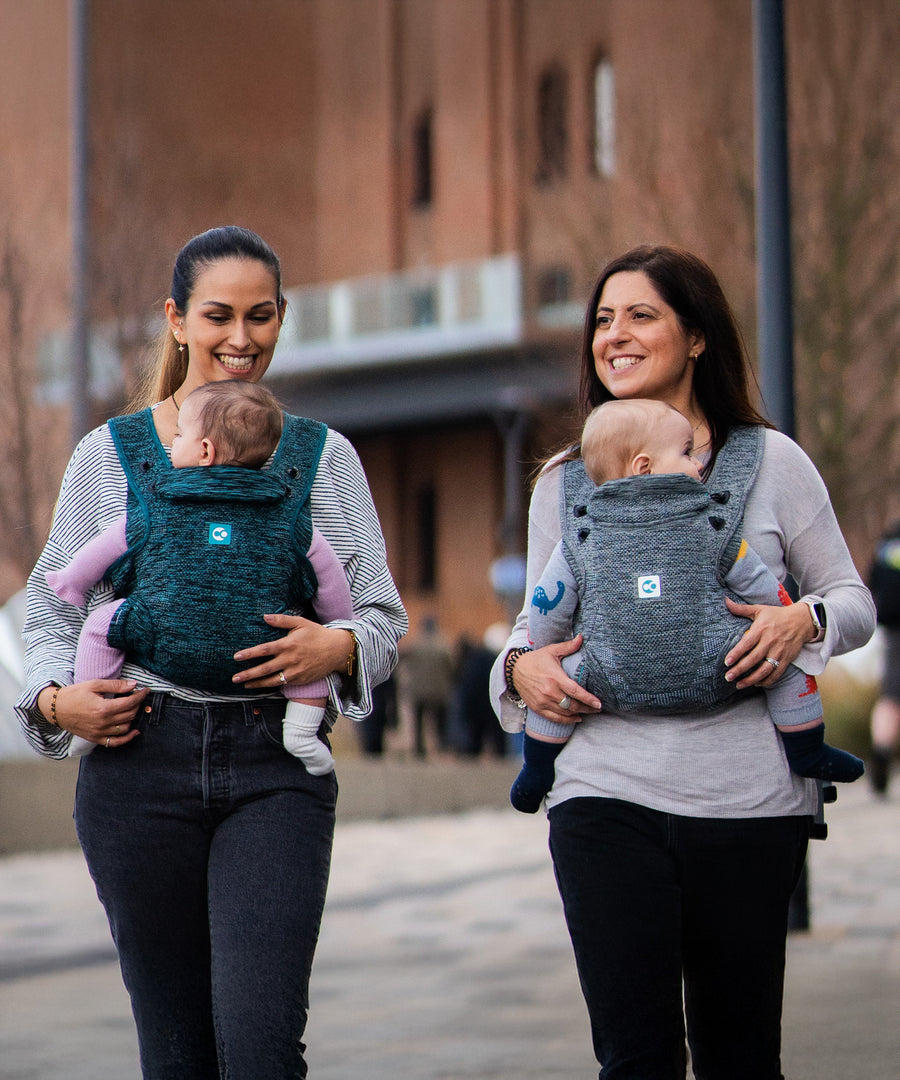 Two adults carrying a child in the Carifit+ Fabric Baby Carrier in Cool Grey and Cool Green with a town/city scape scene behind them