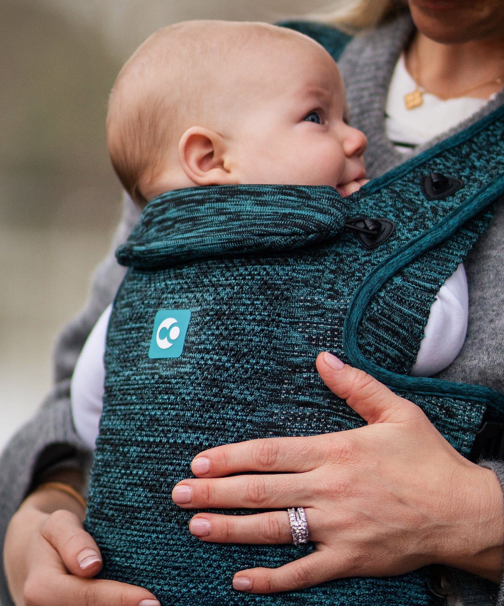 A child facing inwards whilst being carried in the Carifit+ Baby Carrier - Cool Green. The image offers a closer look at the material detailing and the removable head and neck support