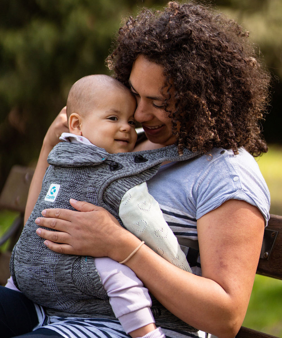 An adult carrying a child inside the Carifit+ Fabric Baby Carrier in Grey. The child is inwards facing towards the adult