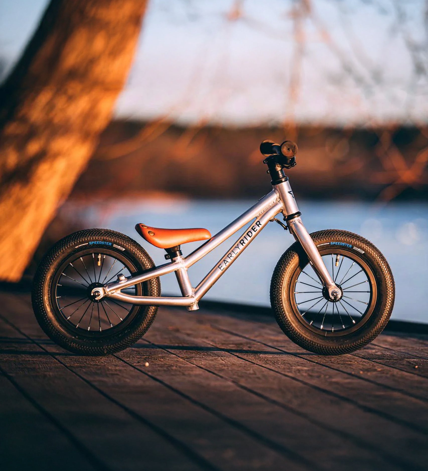 The Early Rider Charger Balance Bike - Violet Haze is stood on a dark wood decking, with a calming lake and tree background in the setting sun