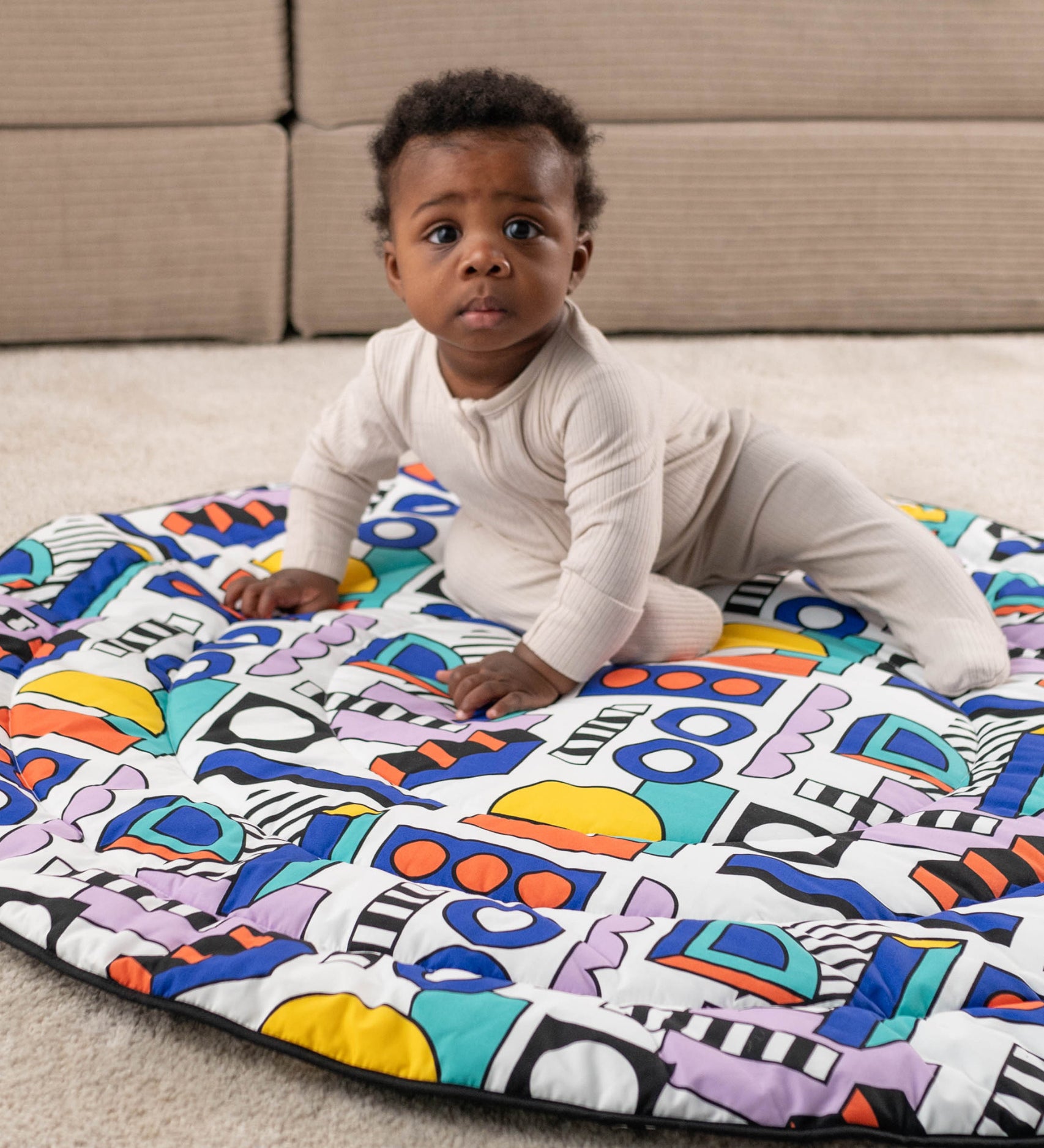 A child happily sat on the Etta Loves x Camille Walala Reversible Playmat, exploring the colourful shape print