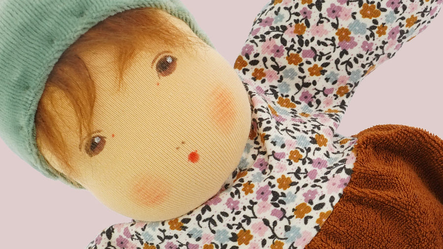 Close up of a beautiful nanchen Waldorf doll, focusing on the face. The doll wears a light green pixie hat, with mohair fringe peeking out, painted on natural expressions including brown eyes and  rosy cheeks.  The doll is on a pink background