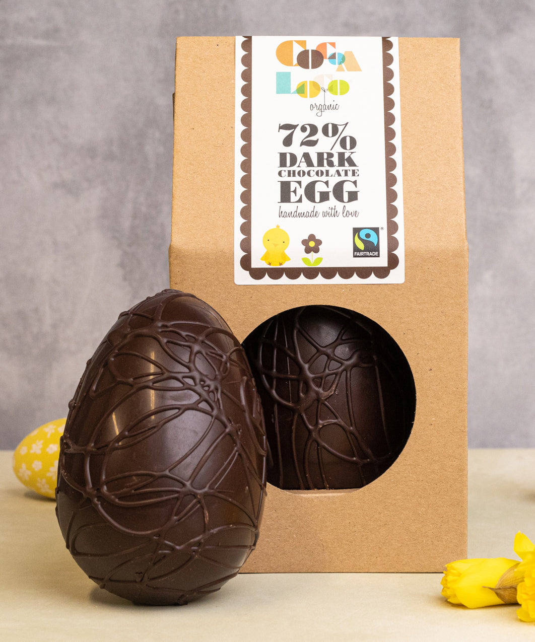 Cocoa Loco organic Fairtrade vegan dark chocolate Easter egg, with chocolate swirl details. Another sits beside it in eco-friendly brown paper packaging, surrounded by bright daffodils and easter eggs.