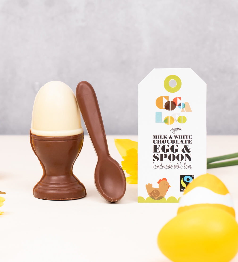 Cocoa Loco Fairtrade organic chocolate egg and spoon set. Solid white chocolate egg in a milk chocolate eggcup with a spoon, designed to look like a boiled egg. Shown with eco packaging, daffodils, and Easter decorations.
