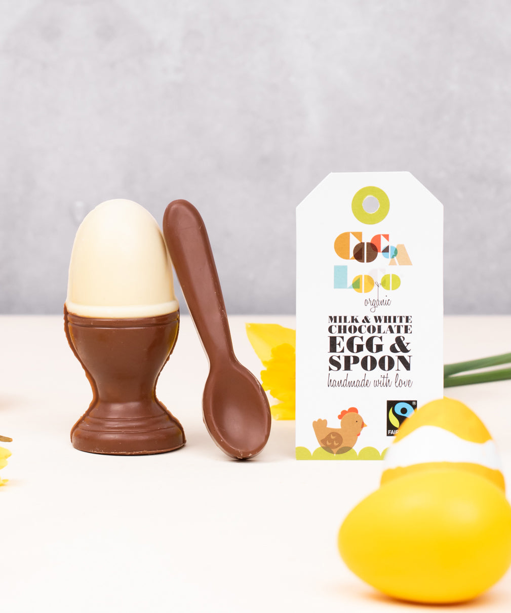 Cocoa Loco Fairtrade organic chocolate egg and spoon set. Solid white chocolate egg in a milk chocolate eggcup with a spoon, designed to look like a boiled egg. Shown with eco packaging, daffodils, and Easter decorations.
