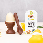 Cocoa Loco Fairtrade organic chocolate egg and spoon set. Solid white chocolate egg in a milk chocolate eggcup with a spoon, designed to look like a boiled egg. Shown with eco packaging, daffodils, and Easter decorations.
