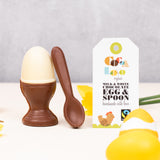 Cocoa Loco Fairtrade organic chocolate egg and spoon set. Solid white chocolate egg in a milk chocolate eggcup with a spoon, designed to look like a boiled egg. Shown with eco packaging, daffodils, and Easter decorations.
