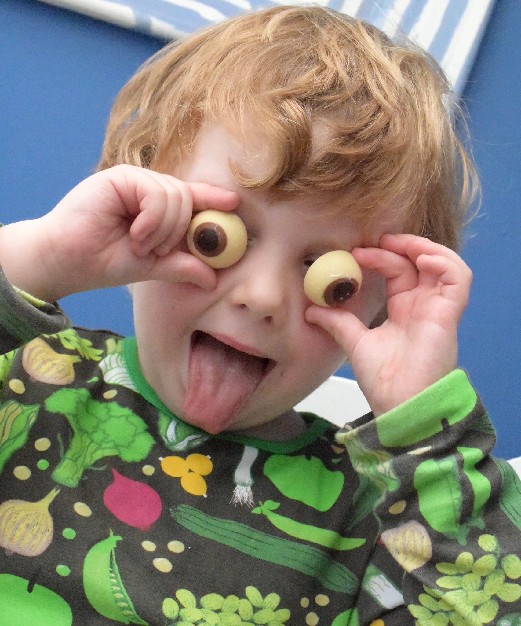 Cocoa Loco chocolate eyeballs being held by a child. The child is sticking his tongue out and is holding the chocolate eyeballs against his eyes. He is wearing a black top with a vegetable print on it.