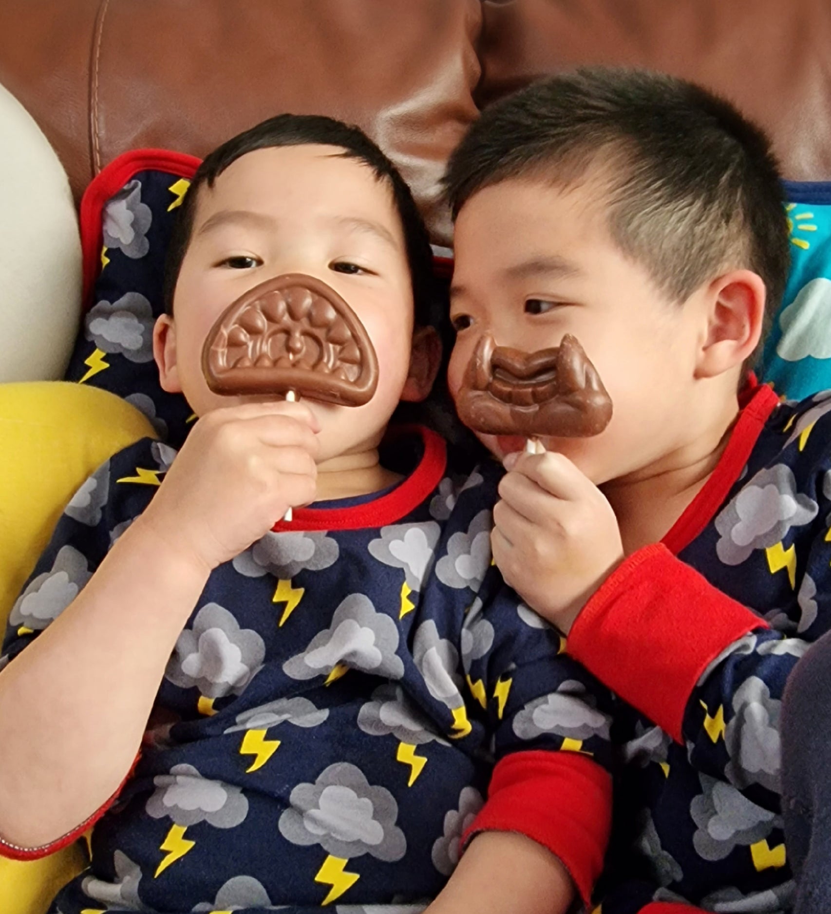 The Cocoa Loco Halloween lollies being held by two playful boys sitting on a sofa. This is a funny picture, they're posing with the lollies over their mouths.