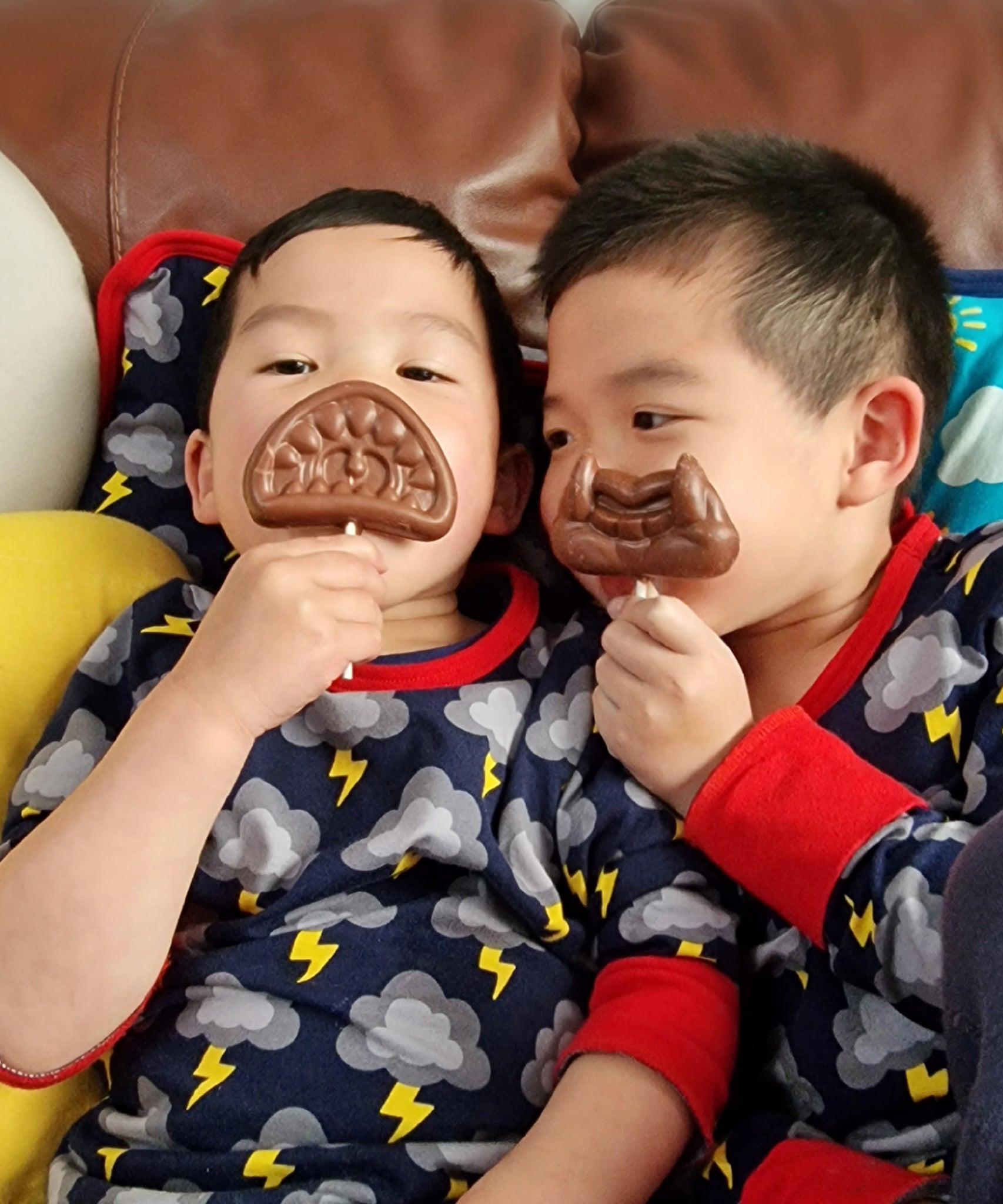 The Cocoa Loco Halloween lollies being held by two playful boys sitting on a sofa. This is a funny picture, they're posing with the lollies over their mouths.