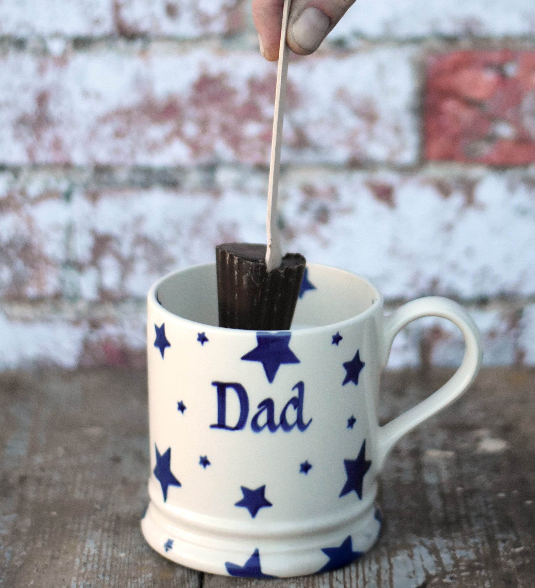 Cocoa Loco hot chocolate spoon in a hand hovering over a white ceramic mug. The mug has a blue star pattern on it and the words dad in the middle.