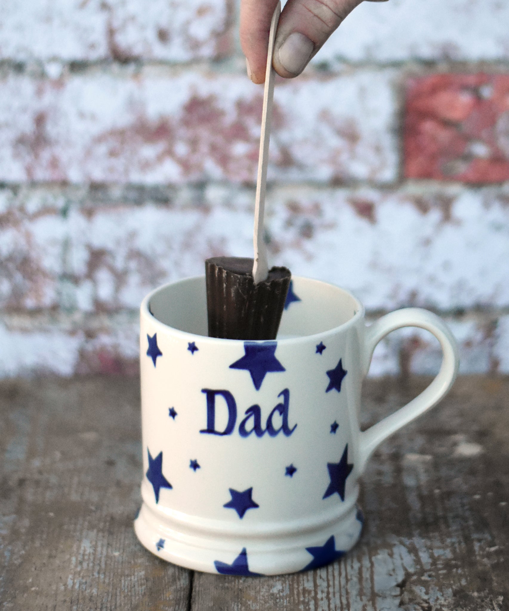 Cocoa Loco hot chocolate spoon in a hand hovering over a white ceramic mug. The mug has a blue star pattern on it and the words dad in the middle.