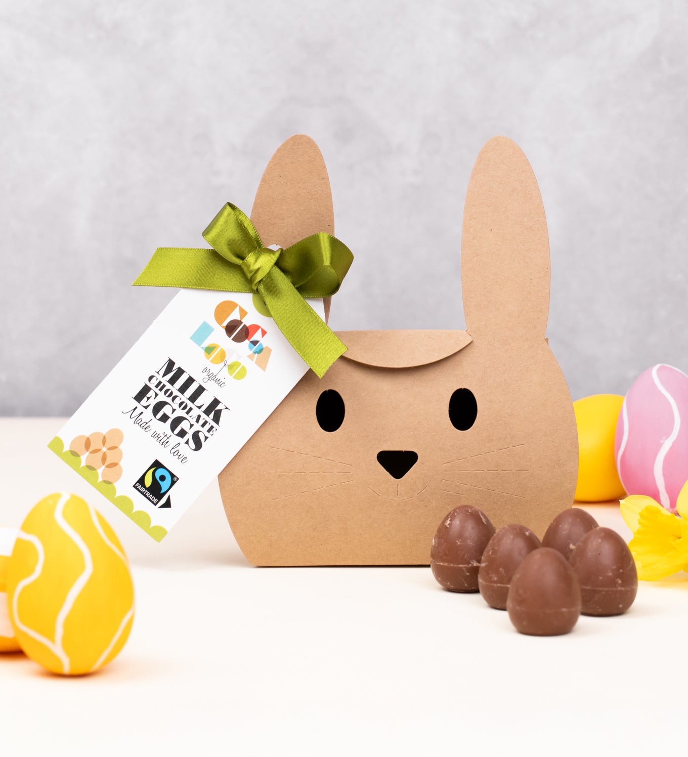 Cocoa Loco Fairtrade organic mini milk chocolate eggs shown with its bunny shaped eco-friendly packaging. Surrounded with colourful mini easter eggs and yellow daffodils.