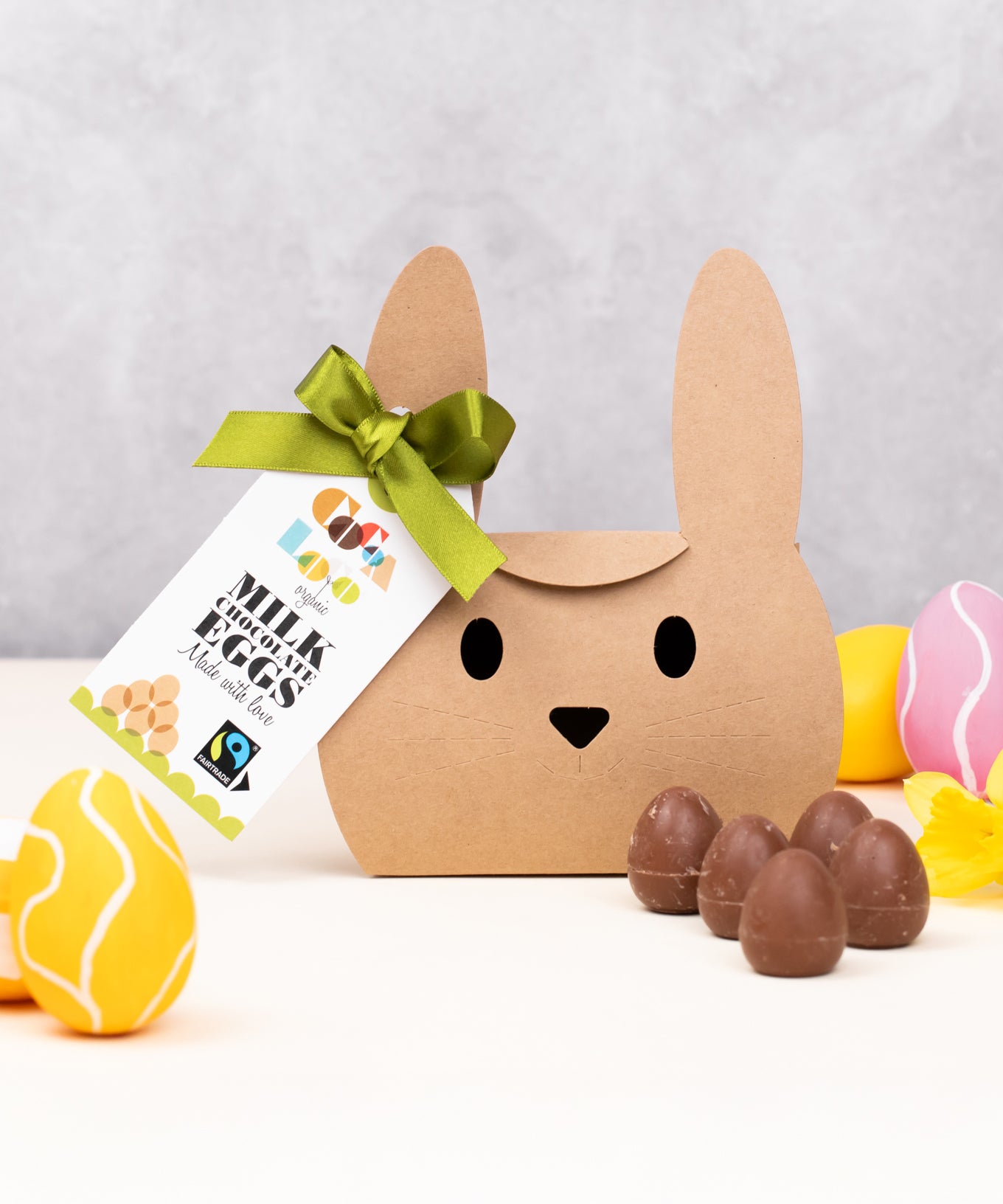 Cocoa Loco Fairtrade organic mini milk chocolate eggs shown with its bunny shaped eco-friendly packaging. Surrounded with colourful mini easter eggs and yellow daffodils.