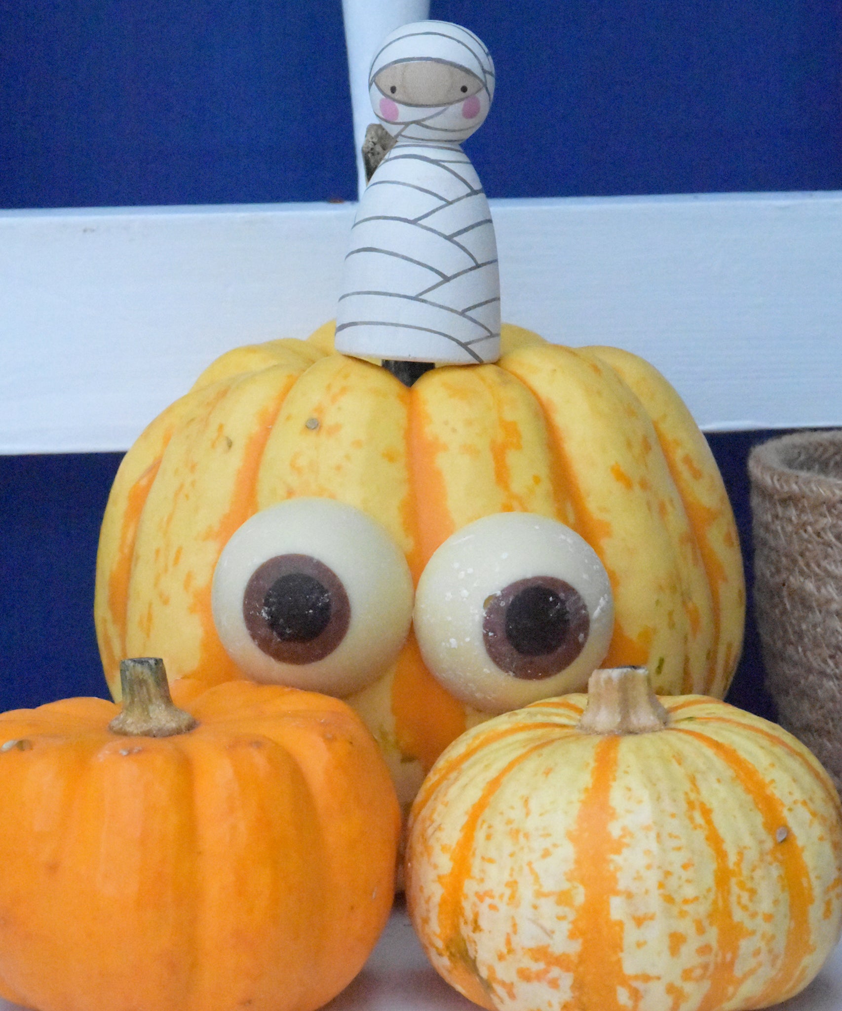 Cocoa Loco chocolate eyeballs stuck on a medium sized orange pumpkin. There is a mummy themed peg doll by Peepul sitting on top of the pumpkin. There are two mini pumpkins in the front of the image.