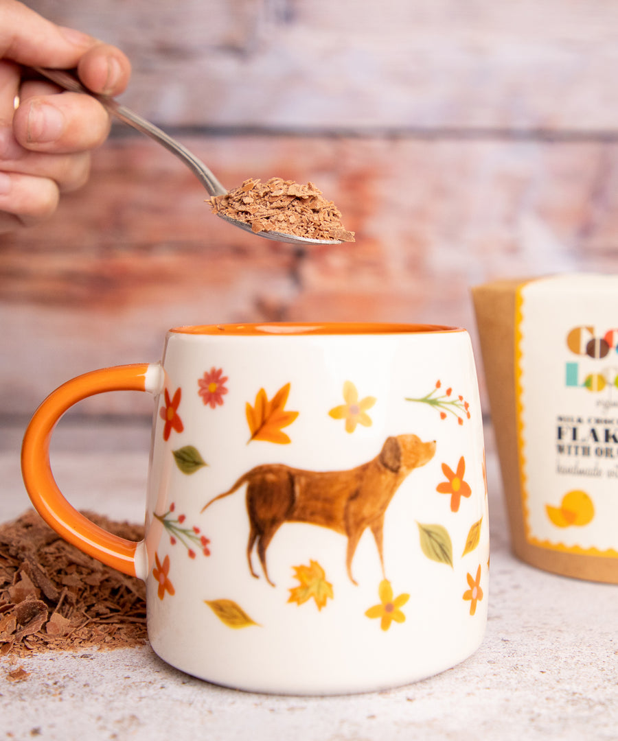 Person putting a spoon of Cocoa Loco Hot Chocolate flakes into an autumnal painted mug.