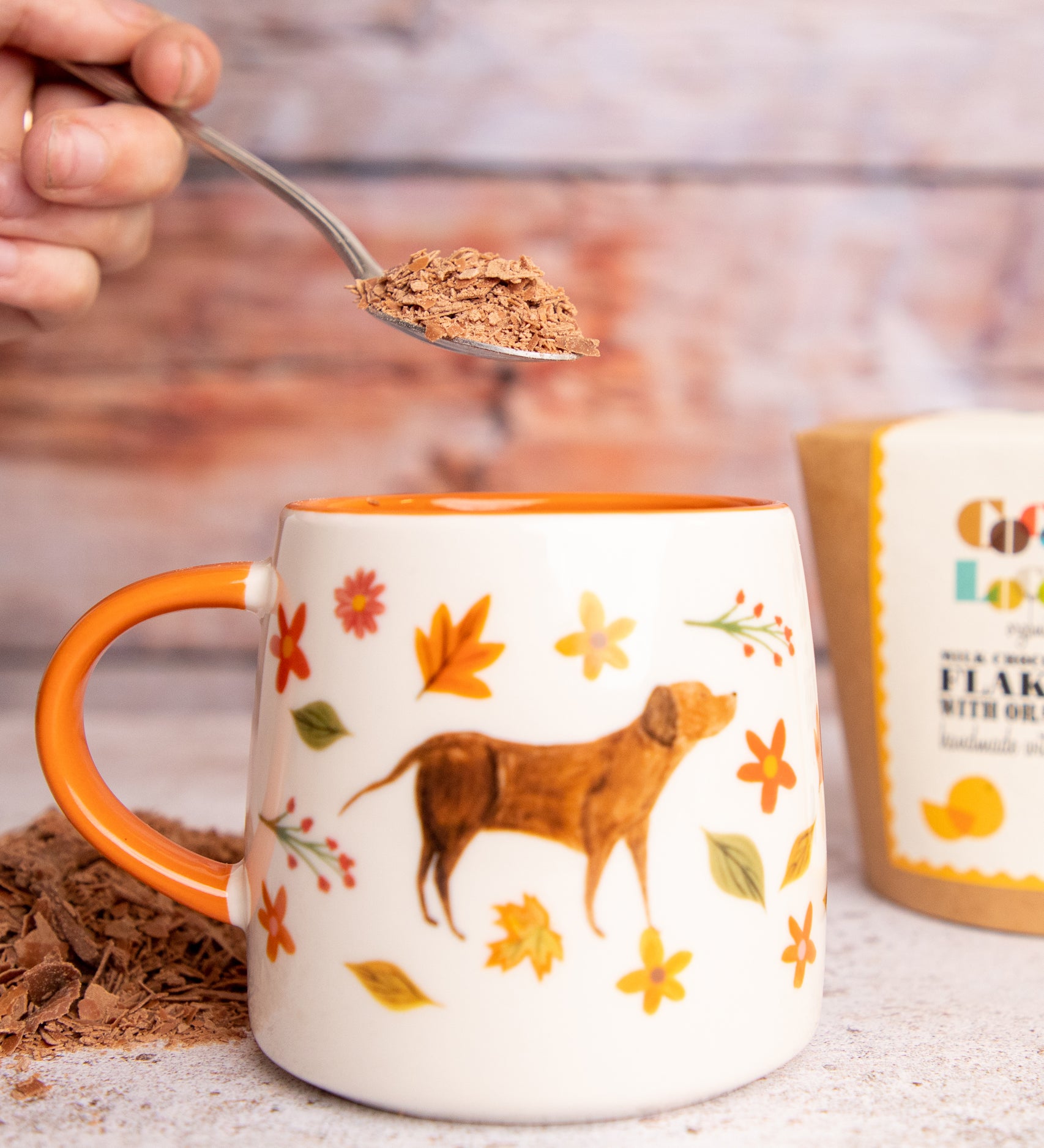 Person putting a spoon of Cocoa Loco Hot Chocolate flakes into an autumnal painted mug.