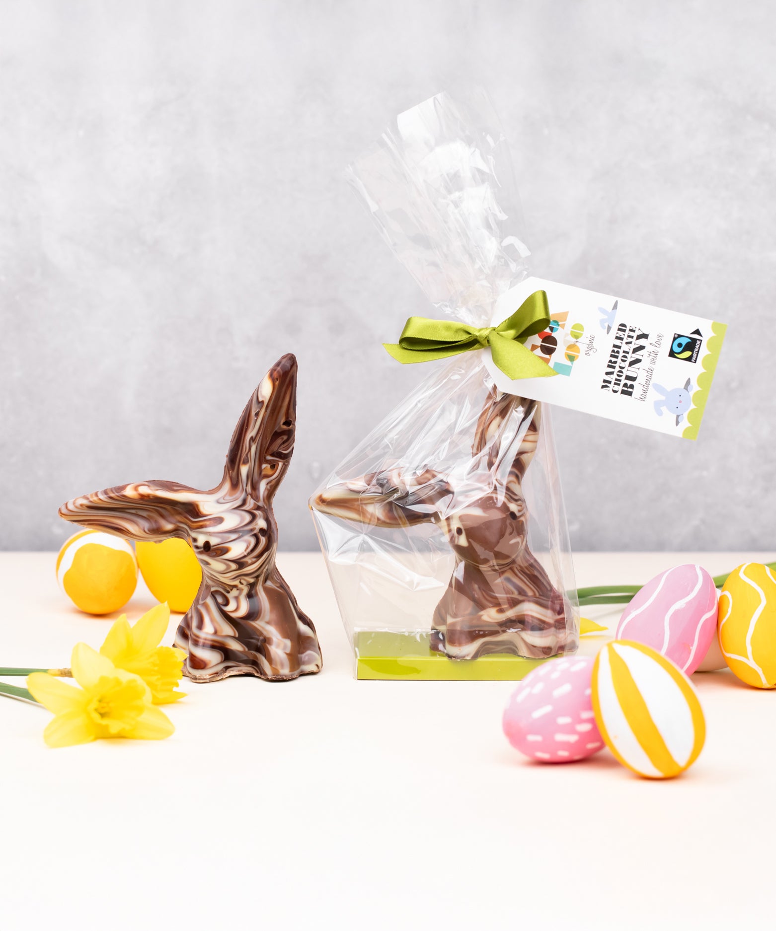 Cocoa Loco organic Fairtrade marbled chocolate rabbit, with one ear up and one down. Another sits beside it in plastic-free packaging, surrounded by colourful mini eggs and daffodils.