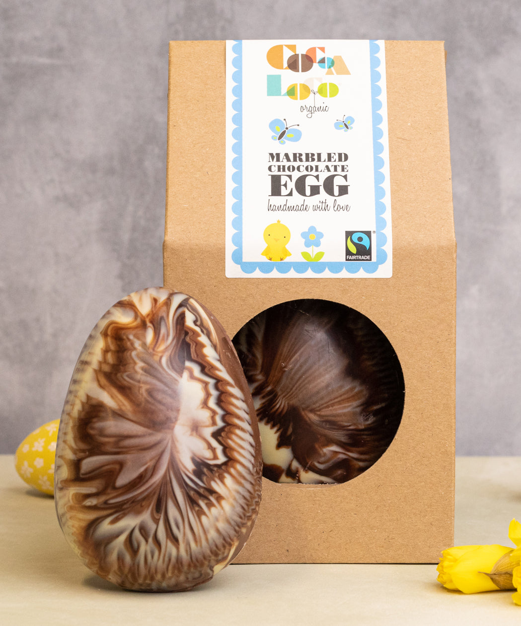 Cocoa Loco organic Fairtrade marbled Easter egg, handmade in the UK with milk, dark, and white chocolate. Another sits beside it in eco-friendly packaging, surrounded by mini eggs and daffodils.