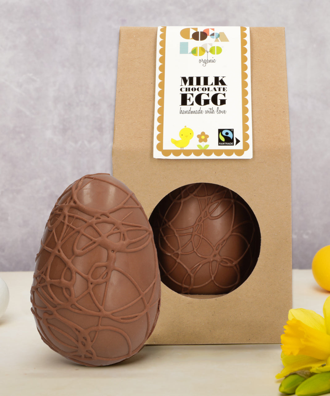 Cocoa Loco organic Fairtrade milk chocolate Easter egg, elegantly swirled with chocolate detail. Another sits beside it in eco-friendly brown paper packaging, surrounded by bright daffodils.
