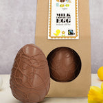 Cocoa Loco organic Fairtrade milk chocolate Easter egg, elegantly swirled with chocolate detail. Another sits beside it in eco-friendly brown paper packaging, surrounded by bright daffodils.
