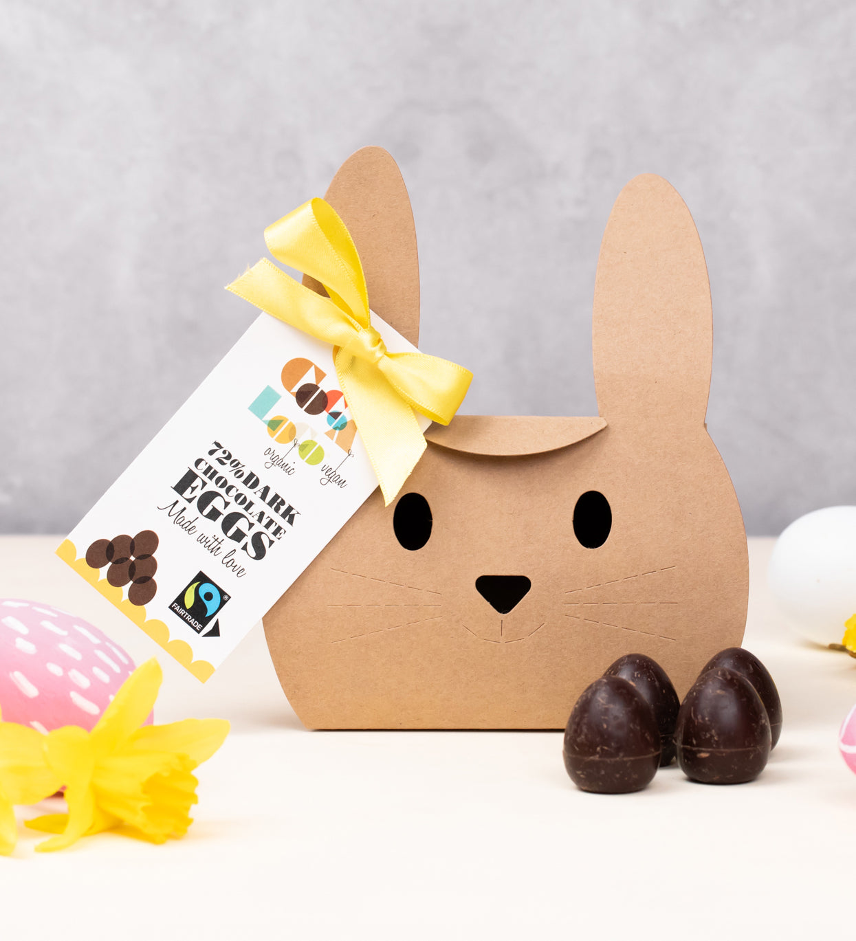 Cocoa Loco Fairtrade organic vegan mini dark chocolate eggs shown with it's bunny shaped eco-friendly packaging. Surrounded with colourful mini easter eggs and bright daffodils.