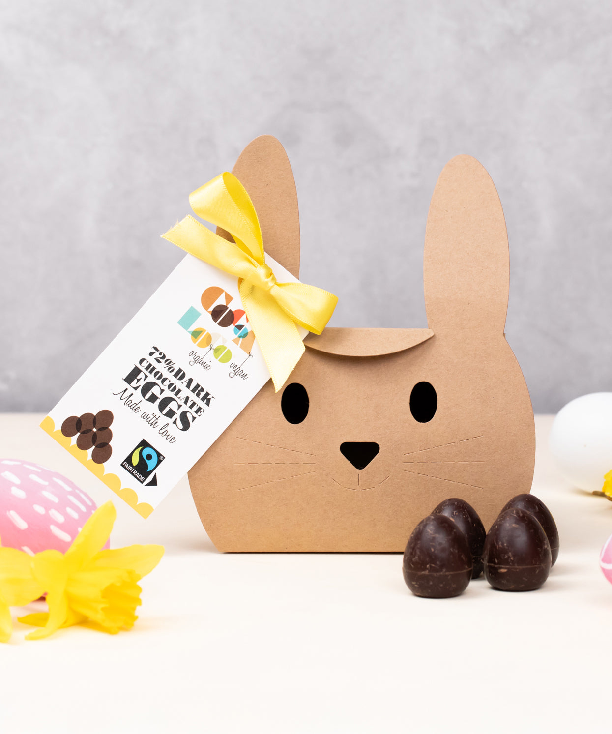 Cocoa Loco Fairtrade organic vegan mini dark chocolate eggs shown with it's bunny shaped eco-friendly packaging. Surrounded with colourful mini easter eggs and bright daffodils.