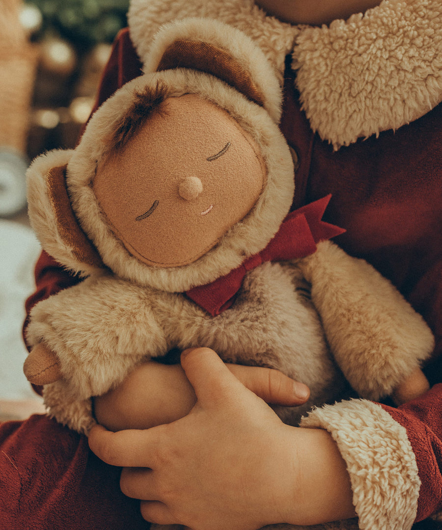 A closer view of the Olli Ella Cozy Dinkums - Teddy Crumble being held by a child's arms. Teddy Crumble as a sleepy smiling face with a cute button nose, and a tuft of brown hair poking out from underneath its hood. The child is wearing a red Santa outfit with a white fluffy collar.