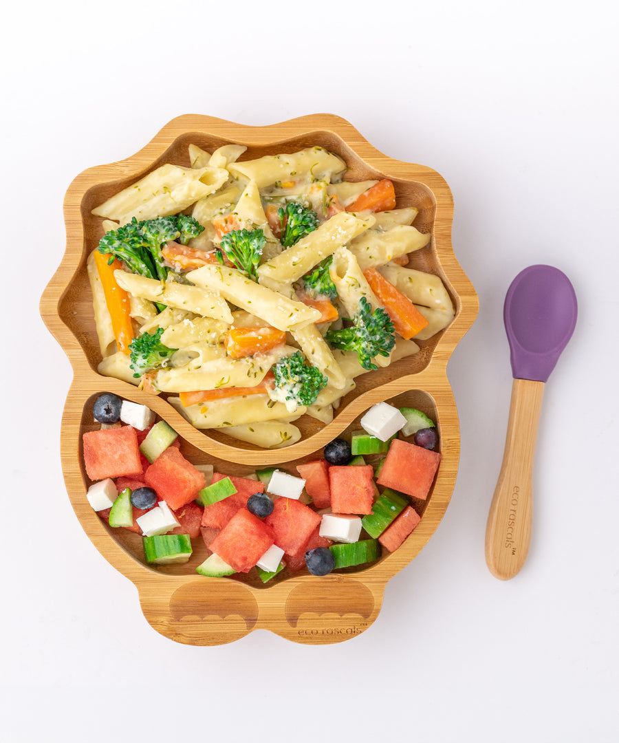 Eco Rascals Dinosaur Bamboo Suction Plate with pasta, carrots and broccoli and a watermellon side salad, and purple silicone and bamboo baby spoon.