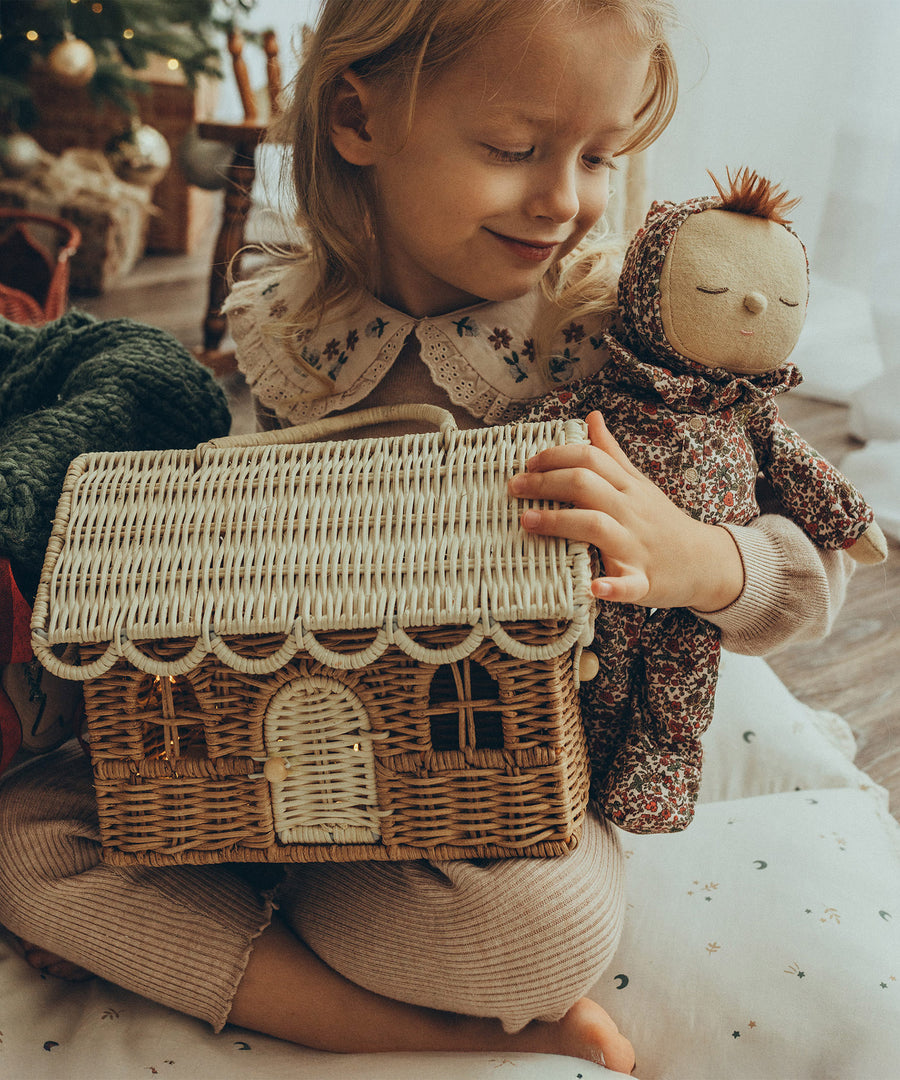 A child sat on a white cushion and is holding Olli Ella Dozy Dinkum Doll Pudding - Garland and the Olli Ella Gingerbread House Basket in their arms. The child is smiling and is looking down at the Dozy Dinkum Doll