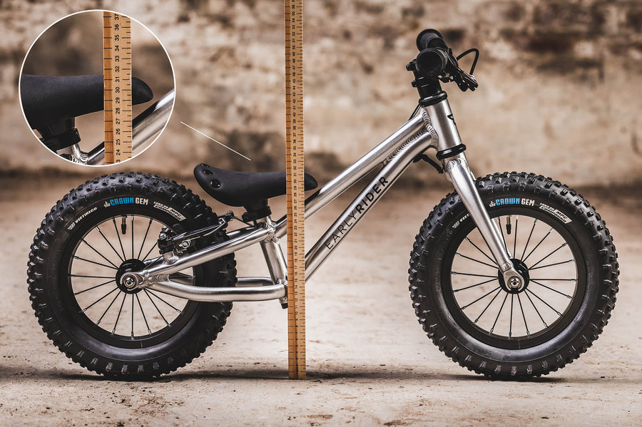 The Early Rider Big Foot Balance Bike stood in front of a light brown wooden ruler, showing the height from the floor to the top of the black seat