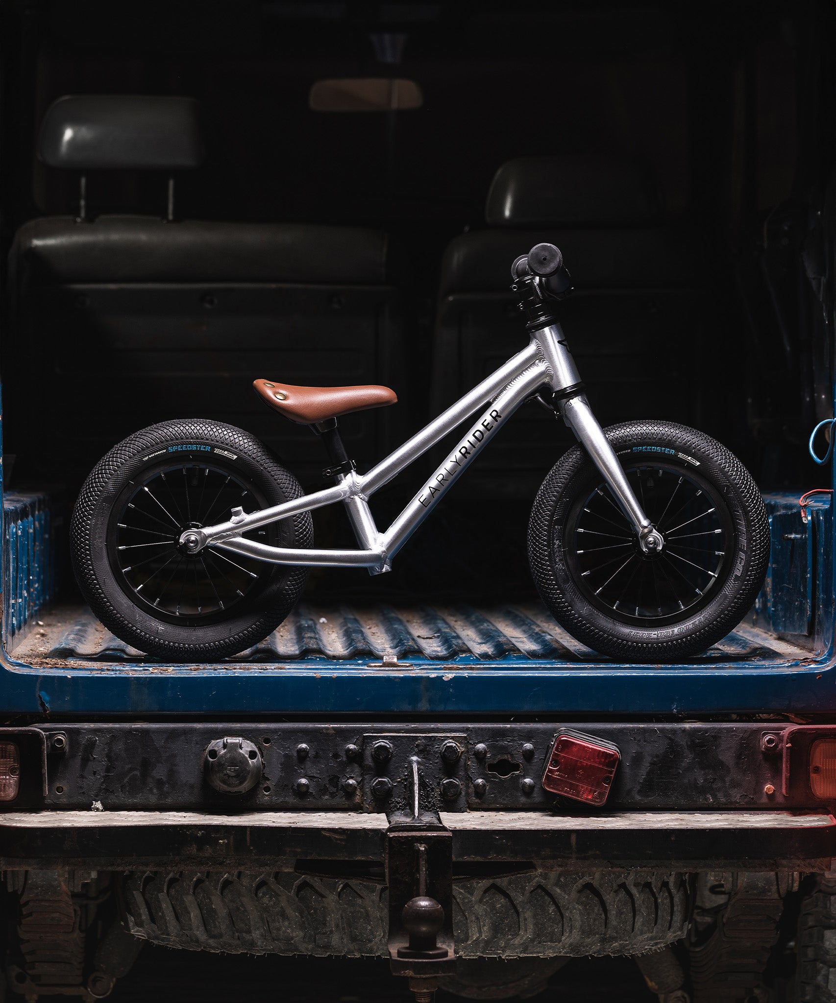 The Early Rider Charger Balance Bike in the back of a blue van. The van is muddy, which represents the hardiness of the Early Rider bikes, and their ability to be used on different terrains.