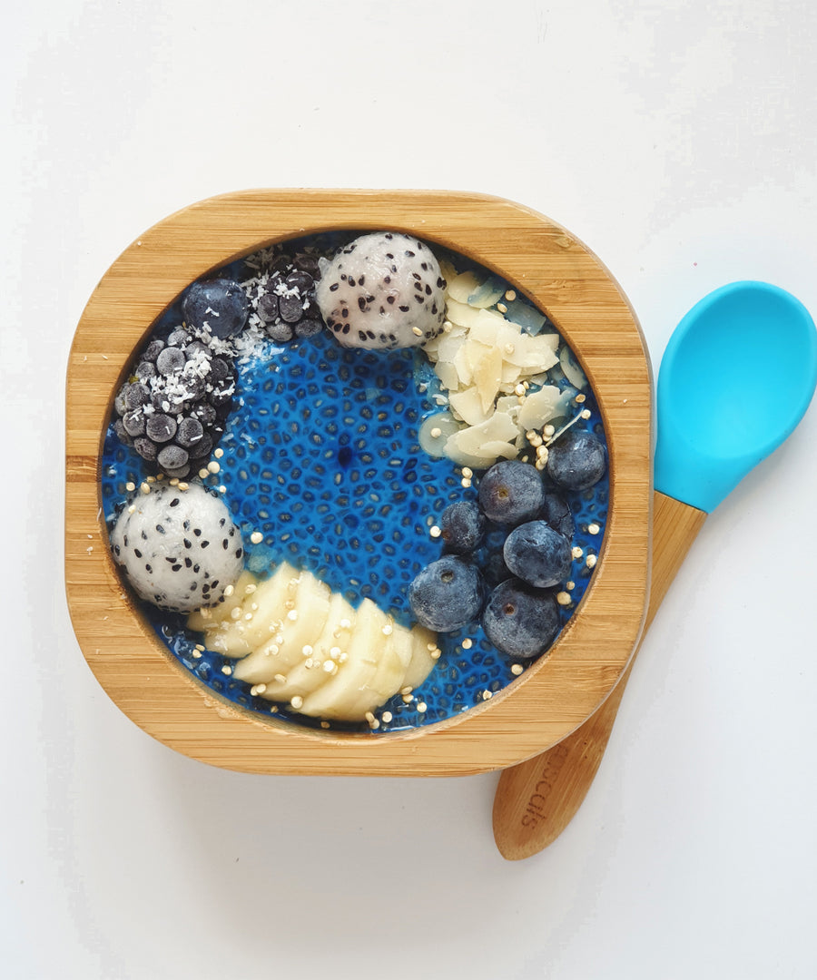 A bamboo baby weaning bowl with blue silicone spoon. The bowl is filled with a blue chia pudding, blueberries, and banana slices. 