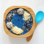 A bamboo baby weaning bowl with blue silicone spoon. The bowl is filled with a blue chia pudding, blueberries, and banana slices. 