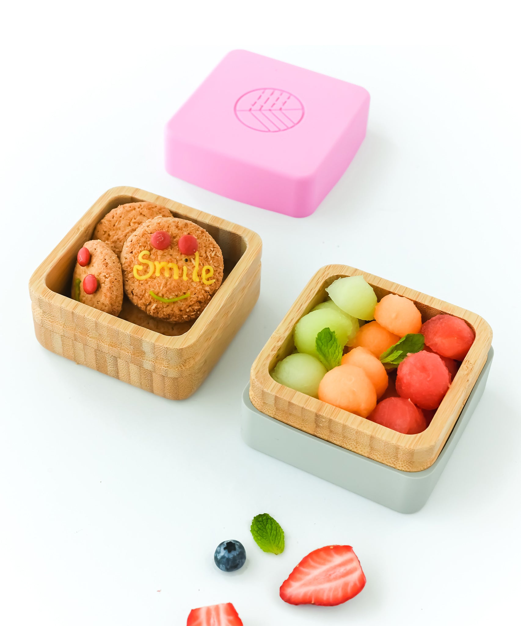 Two open bamboo snack pots, one holding biscuits, the other melon balls with the pink lid above and white lid beneath the pot. 