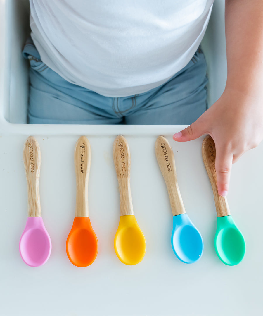 5 bamboo baby weaning spoons lined up on a high chair with a toddler choosing the colour they would like. 