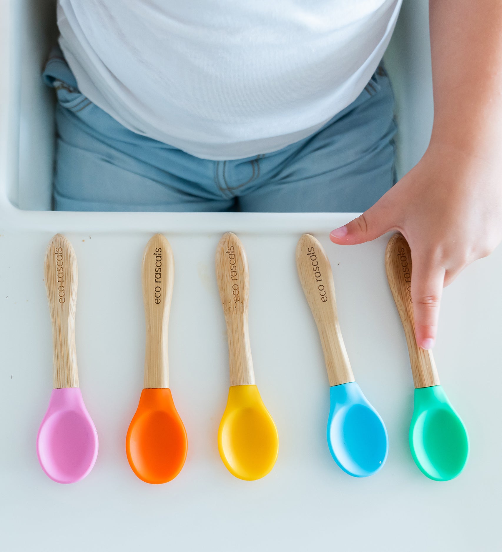 5 bamboo baby weaning spoons lined up on a high chair with a toddler choosing the colour they would like. 