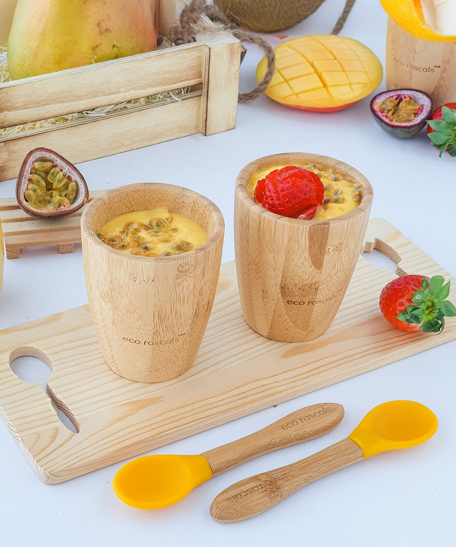 Yellow passionfruit smoothies in two bamboo baby cups on a wooden board and two yellow silicone and bamboo baby spoons, decorated with strawberrys.